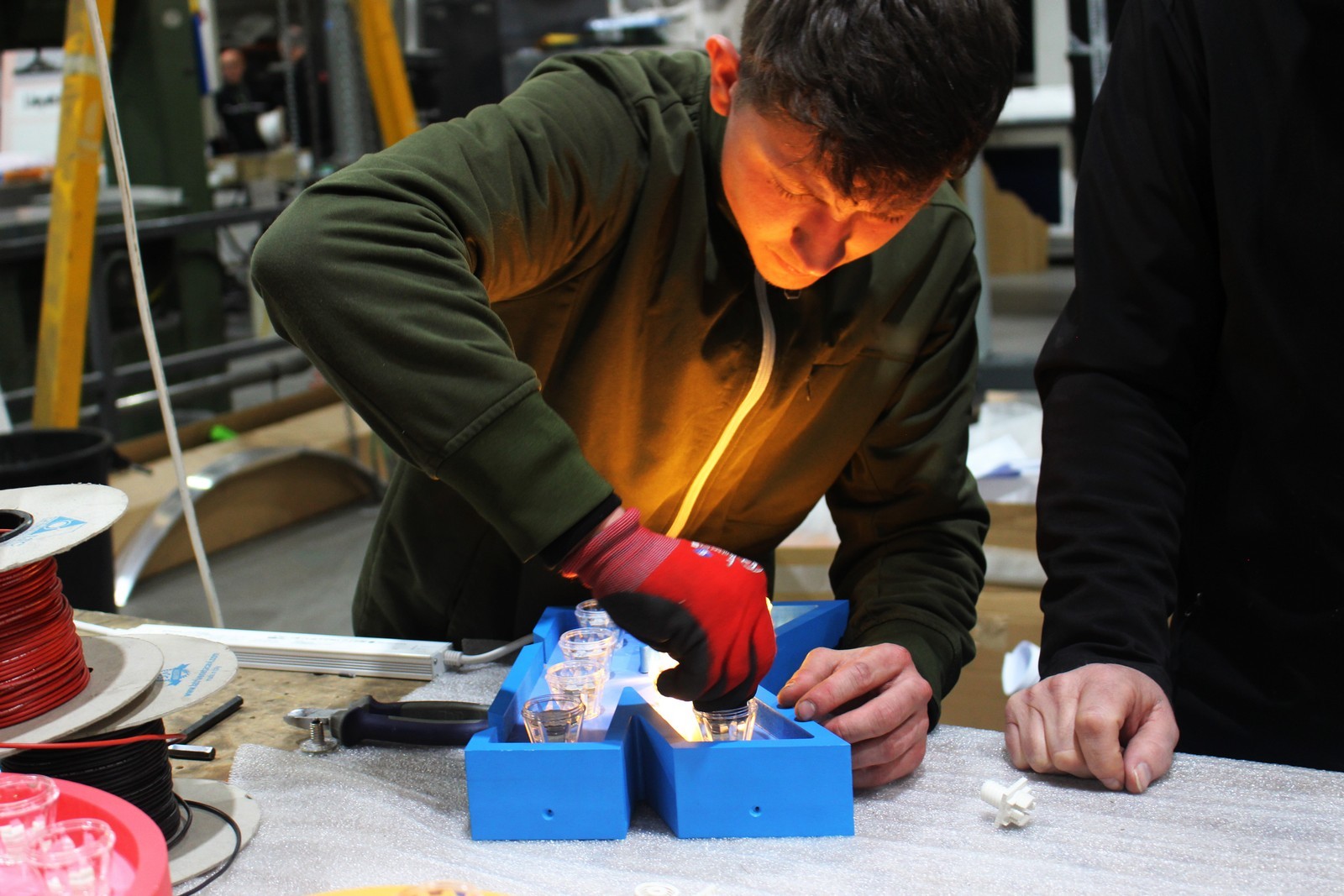 A person in a green jacket works on a blue mold with glowing lights.