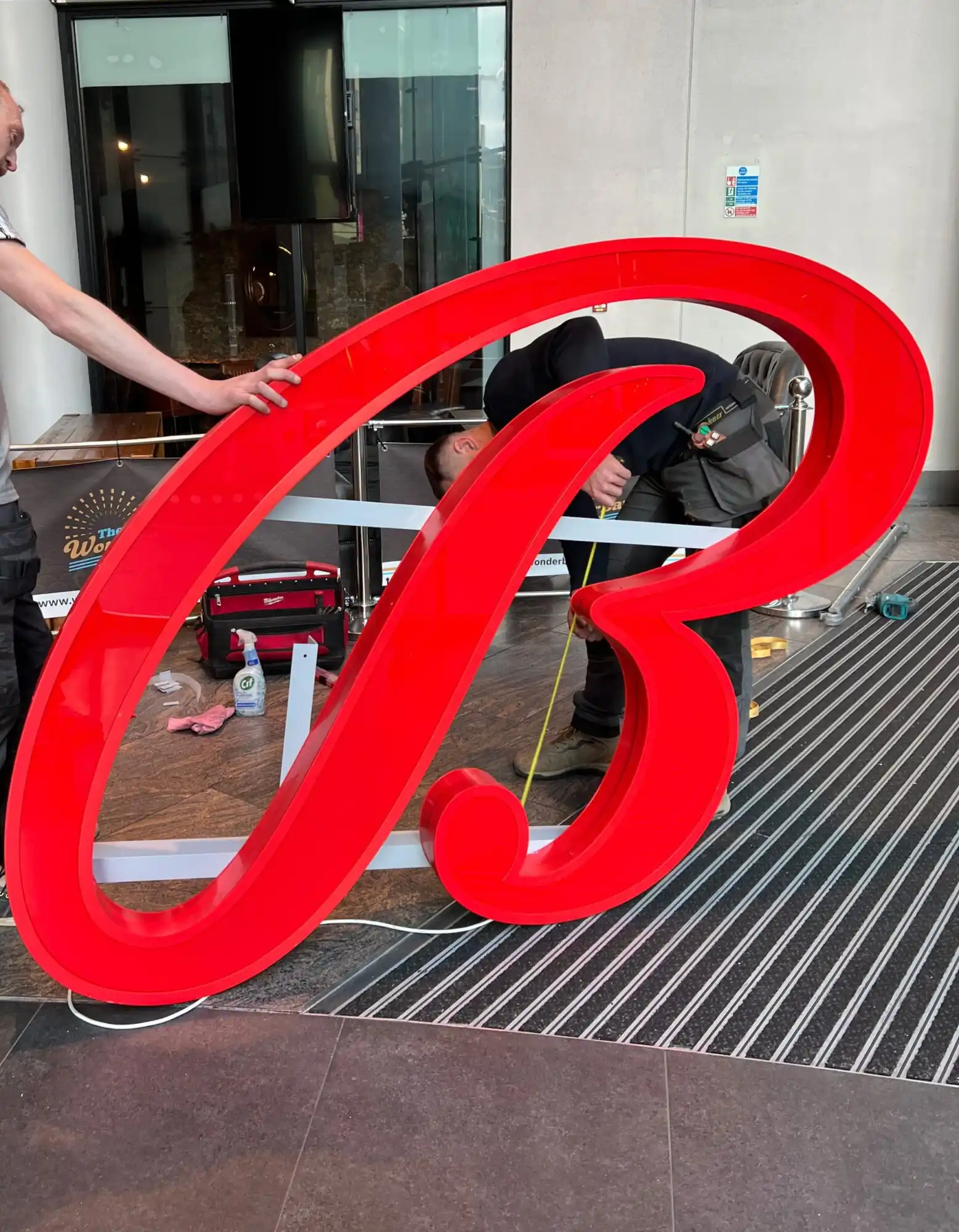Two people hold a large, red, illuminated letter "B" inside a modern building.