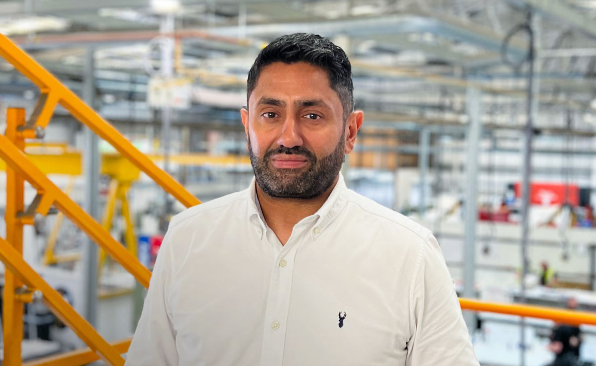 Man in a white shirt with a small emblem stands in an industrial setting, featuring orange railings and machinery in the background, suggesting a factory or manufacturing environment.