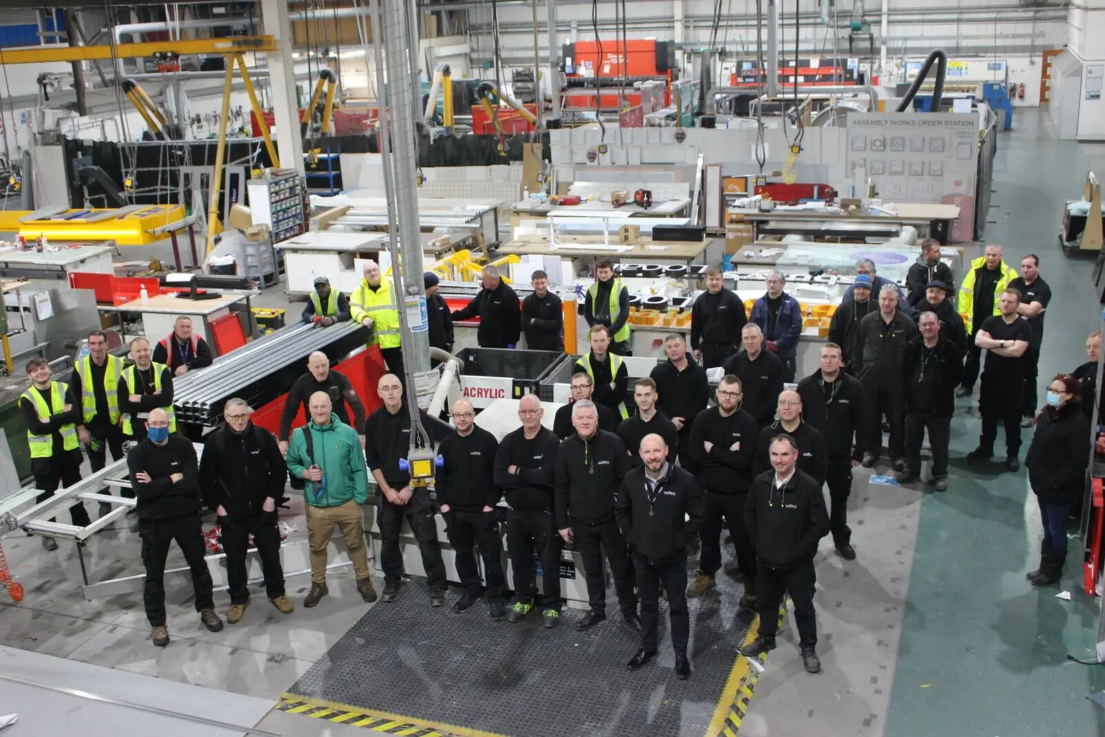 Large group of factory workers in uniform standing in an industrial warehouse with machinery and equipment. The setting includes various metal and electronic components, safety signs, and high-visibility jackets. The environment indicates a manufacturing or production facility.