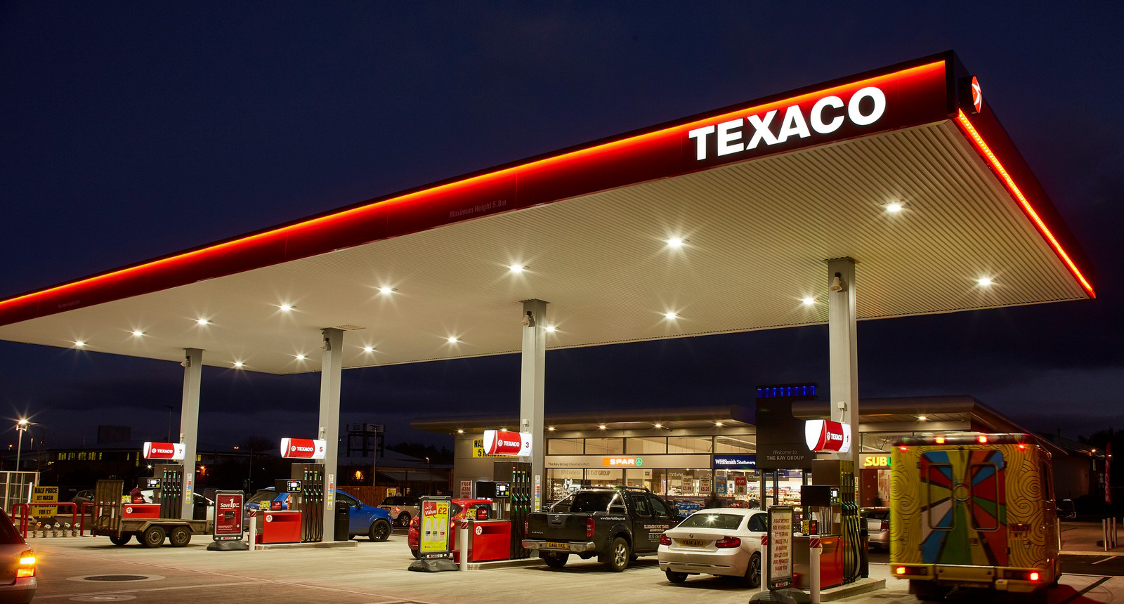 Texaco gas station at night with illuminated canopy and several cars parked at pumps.