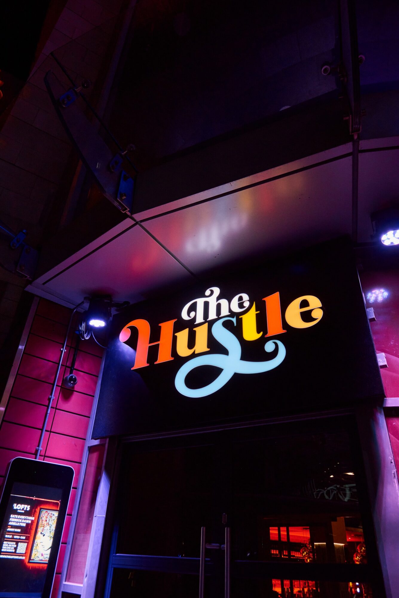 Illuminated sign reading "The Hustle" above a building entrance at night.