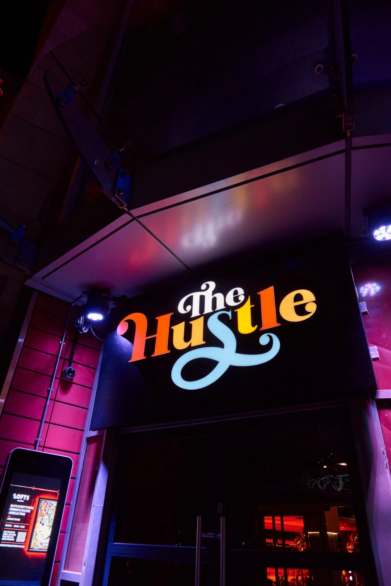 Illuminated sign reading "The Hustle" above a building entrance at night.